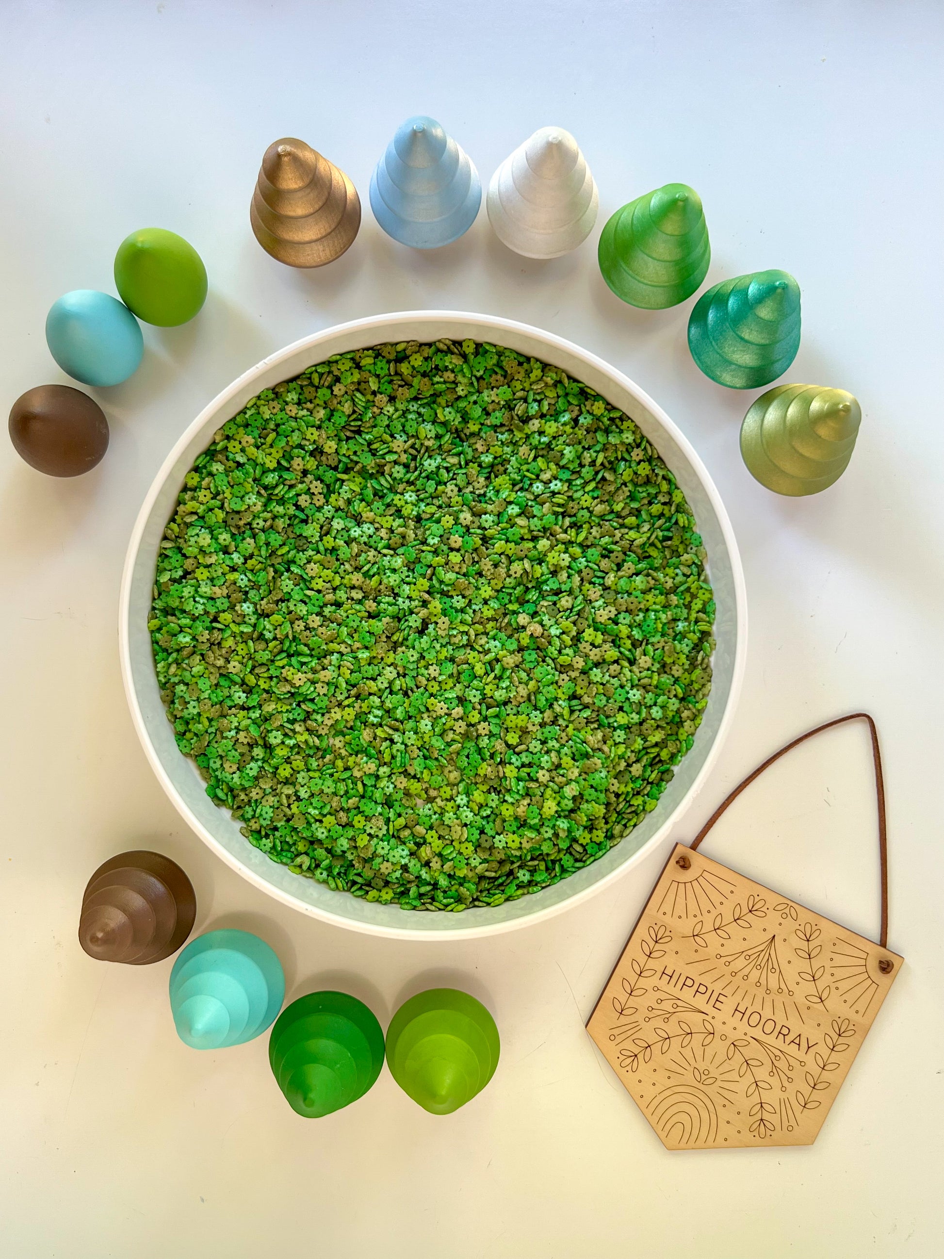White bowl filled with green leaf inspired sensory filler surrounded by colorful wooden trees and a wooden hippie hooray card on a light background