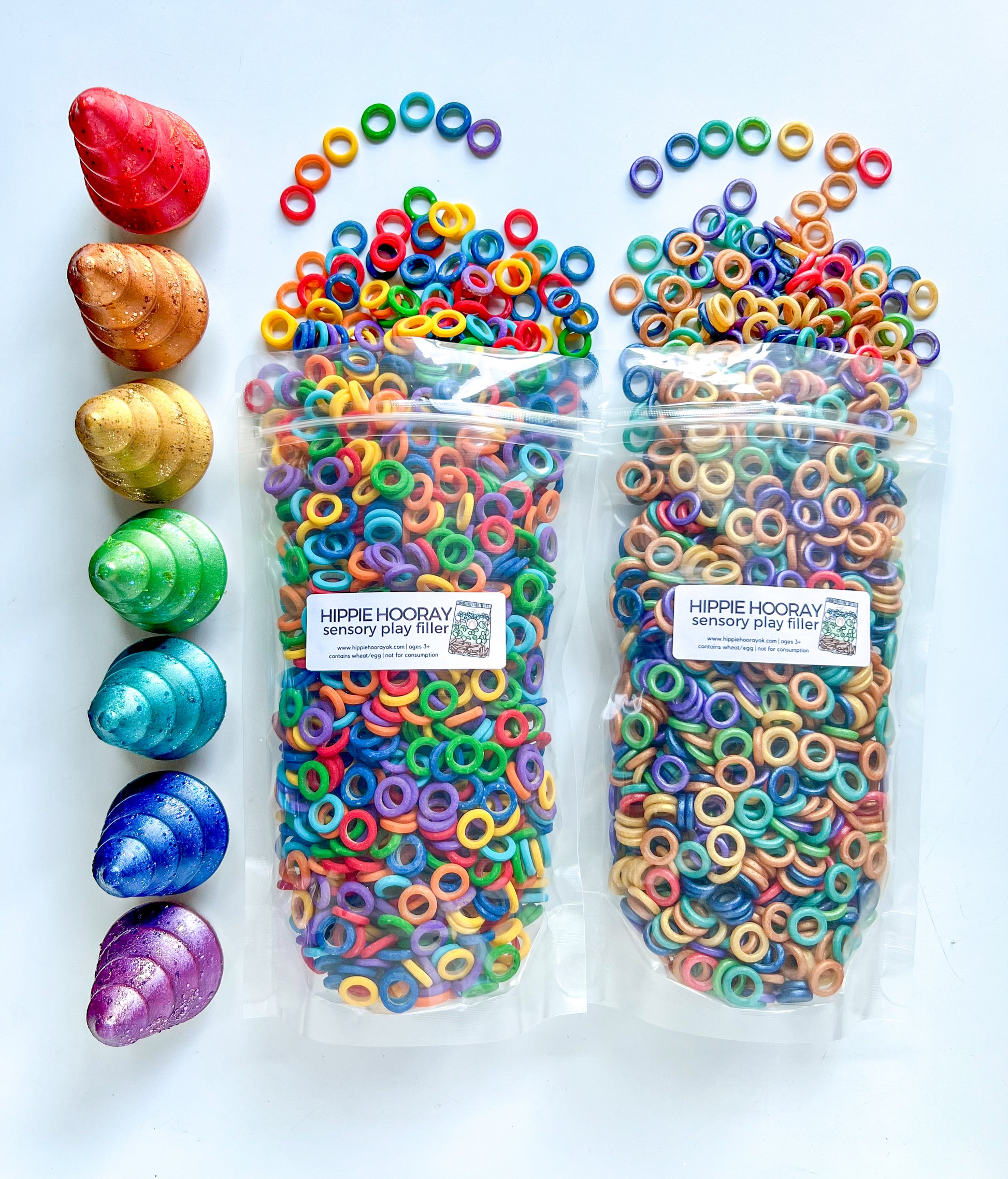 Packaged sensory bin fillers with various shapes and colors designed for sensory play, displayed alongside loose rings and pasta in a rainbow pattern. Hippie Hooray rainbow ring pasta and wooden play bowls and trees